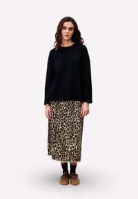 Femme debout portant un haut noir à manches longues, une jupe midi à imprimé léopard, des chaussettes noires et des chaussures marron sur un fond uni.