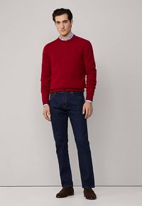 Rode geribde gebreide trui, lichtblauw overhemd met kraag eronder, donkerblauwe spijkerbroek en bruine schoenen. Model poseert rechtop tegen een neutrale achtergrond.