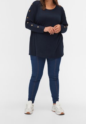 Femme portant un haut bleu marine à manches longues avec des boutons sur les manches, un jean skinny bleu et des baskets blanches, debout avec les mains jointes.