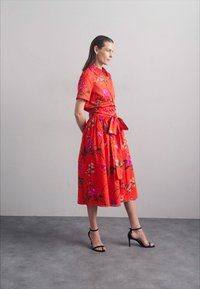 Red floral dress with short sleeves, gathered waist, and knee-length skirt; features pink and green floral patterns. Black high-heeled sandals.