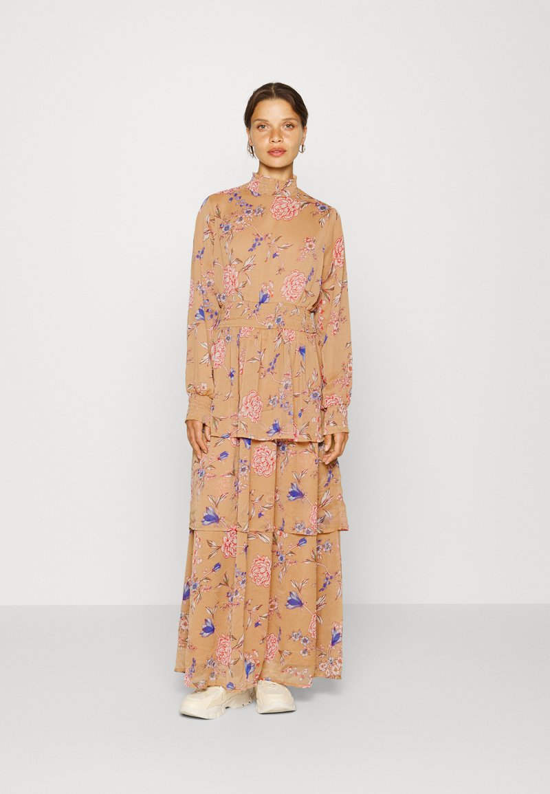 Vestido maxi floral com mangas compridas, gola alta, base bege, padrão floral em rosa e azul, volantes em camadas e caimento descontraído. Usado com sapatos brancos.