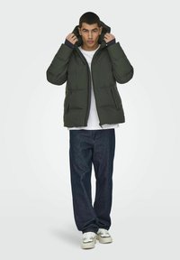 Chaqueta acolchada verde con capucha, frente con cremallera y bolsillos laterales. Llevada sobre una camisa blanca, combinada con jeans oscuros y zapatillas blancas.