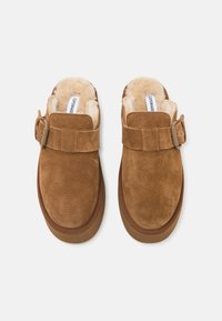 Brown suede slippers with a soft shearling lining, featuring a buckle strap across the instep and a thick rubber outsole. Smooth texture.