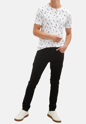 Weißes T-Shirt mit schwarz-weißem Dreiecksmuster, kombiniert mit schwarzen schmal geschnittenen Hosen. Das Modell trägt weiße Sneakers mit tanfarbenen Sohlen.
