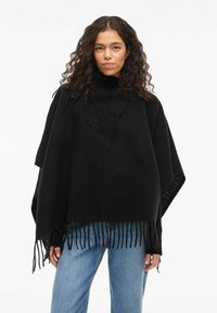Woman with curly hair wearing a black fringed poncho and light blue jeans, standing against a white background.