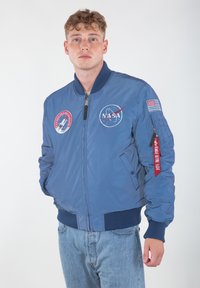 Niebieska kurtka bombowca z naszywkami NASA, ściągaczami przy mankietach i zamkiem błyskawicznym z przodu. Zawiera czerwoną metkę z napisem "Zdejmij przed lotem".