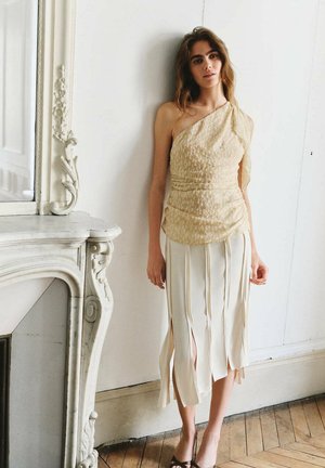 Mujer junto a una chimenea ornamental de mármol blanco, lleva una blusa beige texturizada de un solo hombro y una falda crema con aberturas verticales, con fondo de suelo de madera.