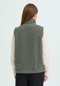 Gilet en polaire de couleur verte atténuée avec un col montant, présentant un tissu texturé et un design sans manches. Couture visible le long du bord inférieur.