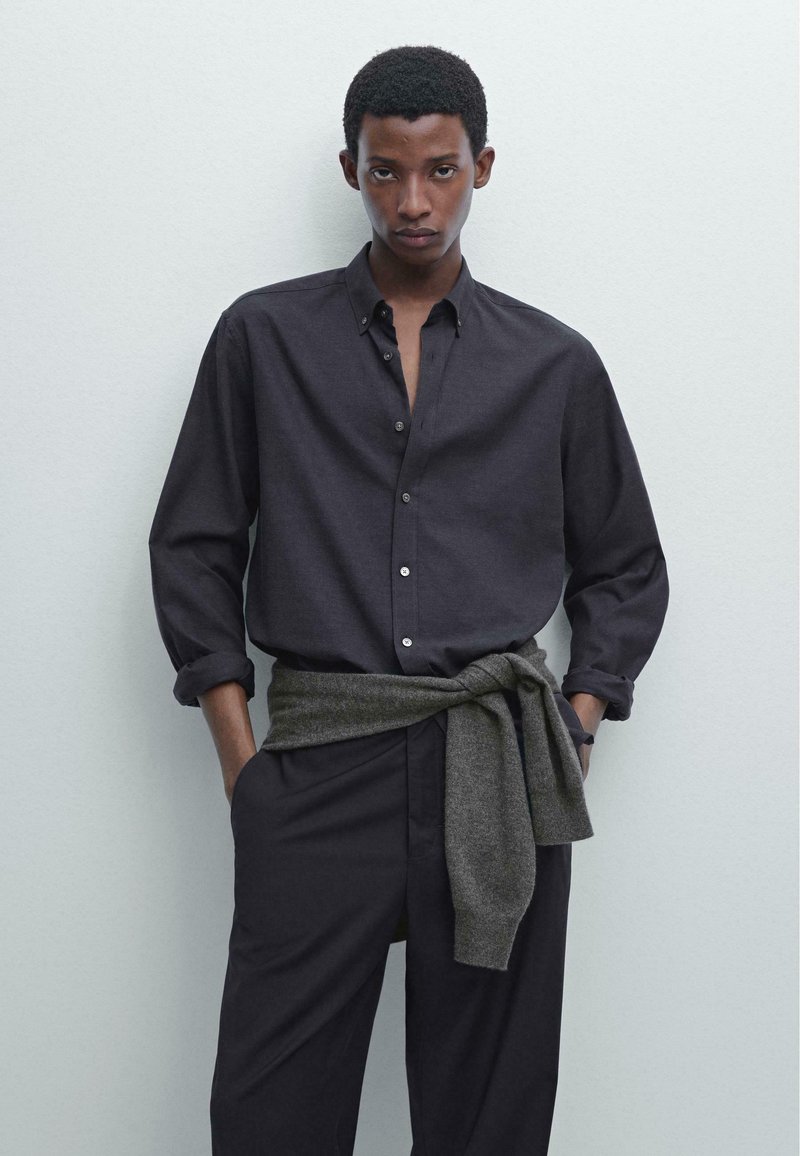 Camicia nera con bottoni e maniche arrotolate, abbinata a pantaloni scuri e un maglione grigio annodato in vita, su uno sfondo chiaro.