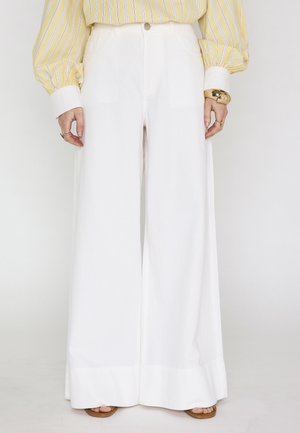 Pantaloni bianchi a gamba larga indossati con una camicetta gialla a righe, bracciale dorato, anelli e sandali aperti.