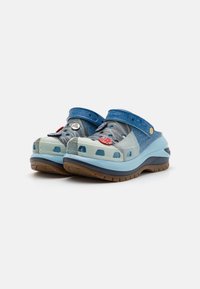 Blå och aqua träskor med en texturerad yta, utrustade med ventilationshål, denimrem och dekorativa nålar. Bruna gummisulor.