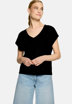 Femme aux cheveux blonds portant un haut noir à manches courtes et col en V ainsi qu'un jean bleu clair, debout, les mains jointes et regardant sur le côté.