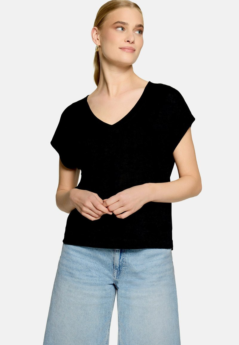 Femme aux cheveux blonds portant un haut noir à manches courtes et col en V ainsi qu'un jean bleu clair, debout, les mains jointes et regardant sur le côté.