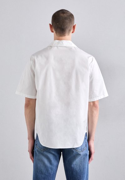 White short-sleeve shirt made of smooth fabric. Features a classic collar and a curved hem. Paired with blue, straight-leg jeans.