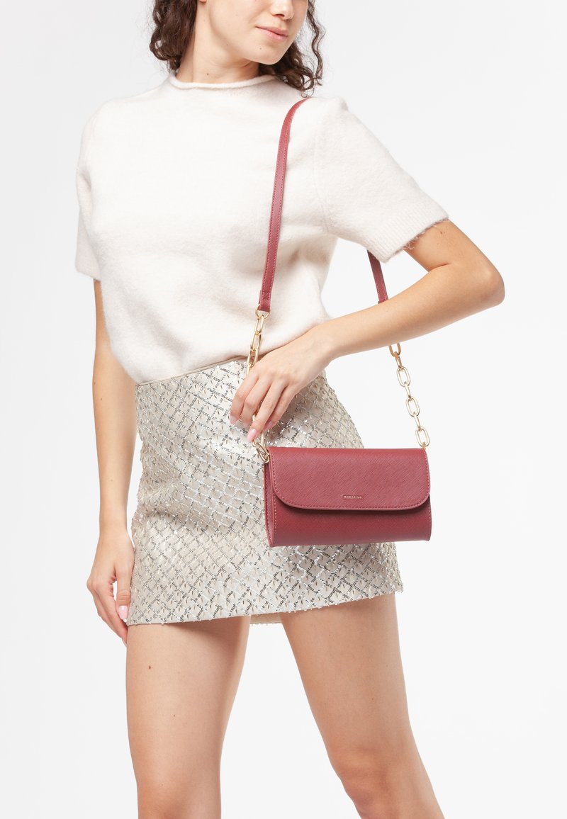 Borsa a tracolla bordeaux con una finitura texture, hardware dorato e una tracolla a catena. Indossata con una mini gonna argento a fantasia.