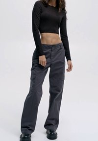 Haut noir à manches longues coupé court, associé à un pantalon cargo gris ample avec poches latérales et fermeture à bouton. Porte des bottes noires.