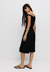Schwarzes Off-Shoulder-Top und knielange schwarze Shorts aus einem weichen Material, kombiniert mit flachen Sandalen mit metallischen Akzenten.