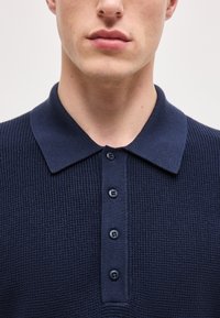 Κοντινό πλάνο ενός ατόμου που φοράει μπλε ναυτικό πλεκτό πόλο πουκάμισο με τέσσερα κουμπιά και μυτερό γιακά.