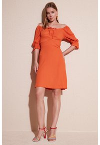 Off-the-shoulder orange dress featuring short puff sleeves and a fitted bodice with ruching. Worn with multi-colored high-heeled sandals.