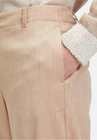 Beige ribbelbroeken met een subtiele glans, voorzien van een zijzak en een gladde textuur. Geaccentueerd met een zachte, lichtgekleurde mouw van een trui.