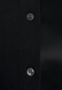 Svart skjorta med knappar i strukturerad, vävd tyg. Har två grå knappar och ett slätt, satinliknande tygpanel på ena sidan.