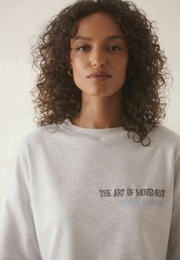 Grauer Pullover mit Rundhals, mit gesticktem Text "THE ART OF MOVEMENT" und "Maître de Ballet" in dunklerem Faden.