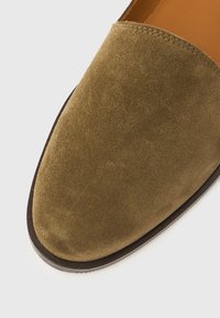 Πράσινο λάδι σουέντ παπούτσι slip-on με στρογγυλεμένη μύτη και σκούρα καφέ ελαστική σόλα.  Ομαλή υφή με ελάχιστες ραφές.