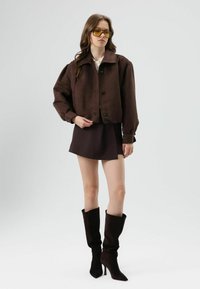 Bruine korte jas met grote mouwen, knoopsluiting, gecombineerd met een bijpassende minirok en hoge, slonzig vallende donkere laarzen. Gele zonnebril inbegrepen.