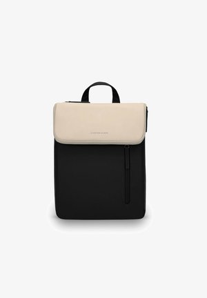 Sac à dos bicolore avec rabat beige, corps noir, poignée supérieure, et poche zippée verticale à l'avant, posé debout sur un fond blanc.