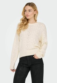Pull beige clair en maille avec une coupe ample, présentant un motif de cœur et des poignets et ourlet côtelés, associé à un pantalon texturé foncé.