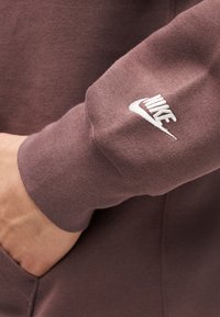 Μανίκι καφέ φούτερ Nike με λευκό κεντημένο λογότυπο, χέρι εν μέρει στην μπροστινή τσέπη.