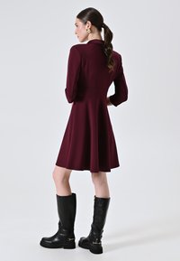Bordeauxfarbenes Kleid mit einem Fit-and-Flare-Design, dreiviertellangen Ärmeln, kombiniert mit knielangen schwarzen Lederstiefeln mit einer robusten Sohle und goldenen Akzenten.