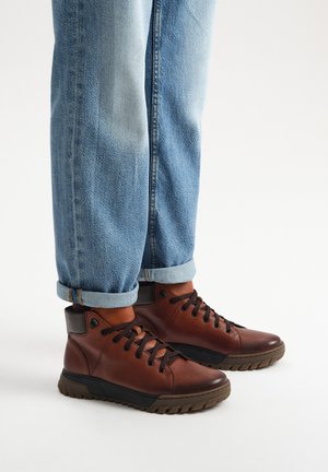 Brązowe skórzane buty za kostkę z czarną gumową podeszwą, zawiązywane, z wyścielanym kołnierzem, w zestawie z podwiniętymi dżinsami.
