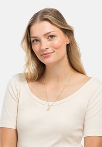 Collana d'oro con pendente circolare, indossata sopra una maglia a coste beige chiaro. La collana presenta un design delicato con anelli texturizzati.