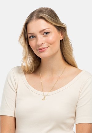 Collier en or avec un pendentif circulaire, porté sur un haut côtelé beige clair. Le collier présente un design délicat avec des maillons texturés.