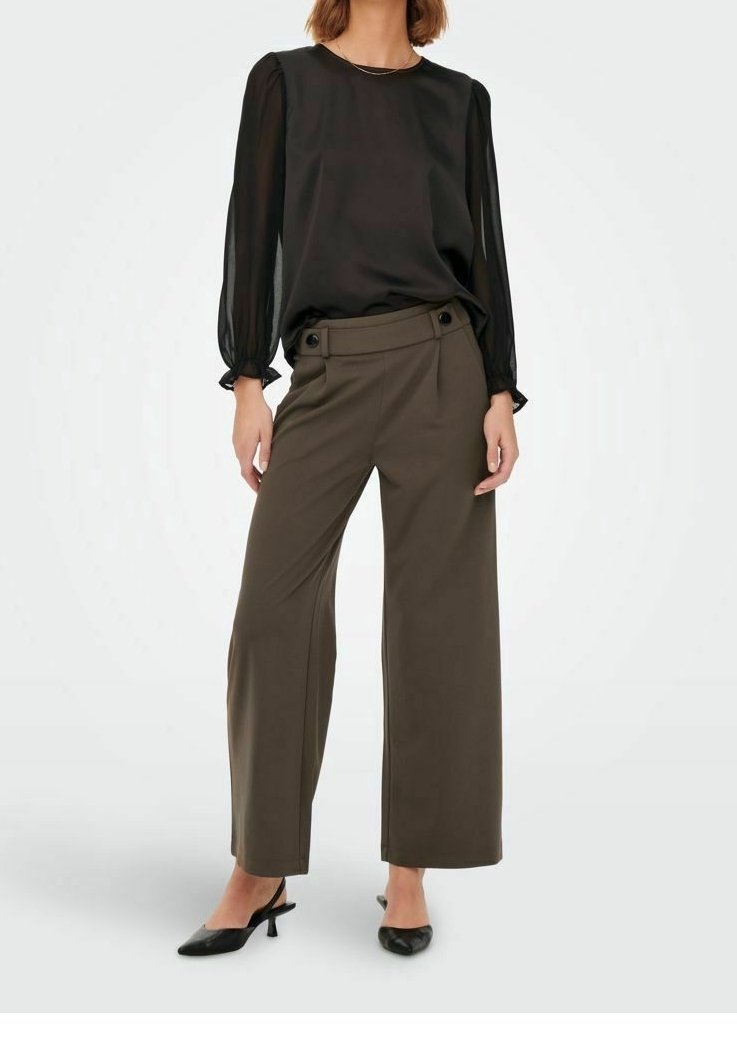 Femme portant un chemisier noir à manches longues, un pantalon large couleur olive avec ceinture boutonnée, et des chaussures slingback noires à talons bas, debout.