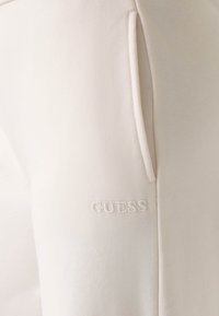 Beżowe spodnie dresowe o miękkiej fakturze, z bocznymi kieszeniami i wyszywanym białym logo "GUESS" blisko dolnego brzegu.