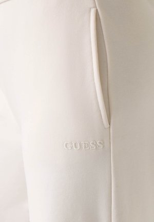 Pantaloni della tuta beige con una texture morbida, dotati di tasche laterali e logo "GUESS" ricamato in bianco vicino all'orlo inferiore.