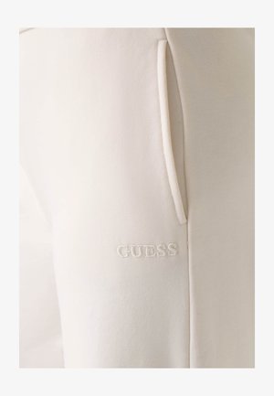 Beige Jogginghose mit weicher Textur, seitlichen Taschen und weiß gesticktem "GUESS"-Logo nahe dem unteren Saum.