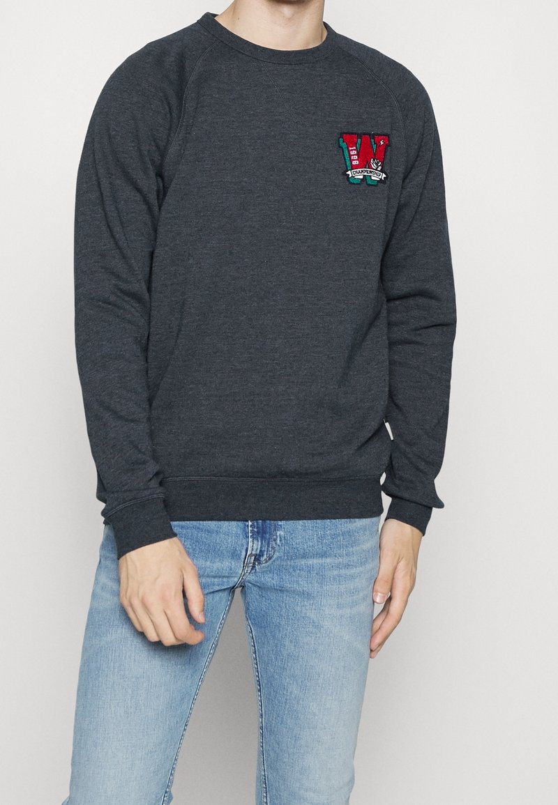 Sudadera gris oscuro de manga larga, con puños acanalados y cuello redondo; presenta un logotipo bordado con una "W" en rojo y teal en el pecho.