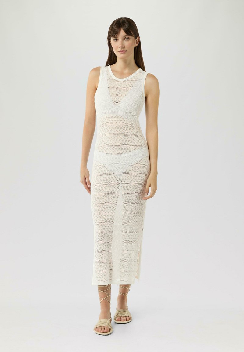 Robe maxi en tricot blanche au design ajusté, présentant un motif géométrique, un col rond et des fentes sur les côtés. Le mannequin porte des sandales neutres.