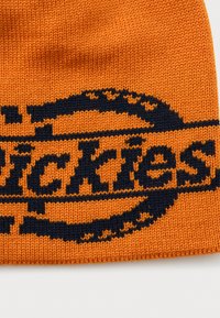 Orange strikket stof med sorte korsknogler og den delvist synlige tekst "Vickies" i fede sorte bogstaver.