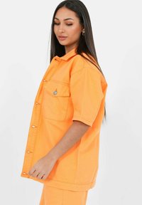 Chaqueta de mezclilla naranja de gran tamaño con mangas cortas, bolsillos frontales y herrajes de metal. Textura suave y ajuste relajado.