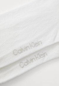 Meias de algodão brancas com textura canelada, apresentando um logótipo prateado "Calvin Klein" perto do punho. O design é simples e sem embellishments.