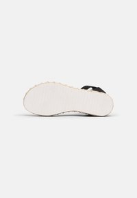 Sandale noire avec une semelle blanche texturée, présentant une bordure en corde de style espadrille. La semelle comporte des rainures verticales pour la traction.