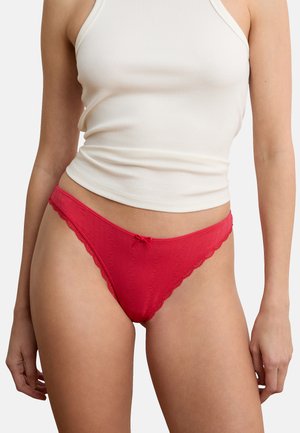 Femme portant un string rouge orné de dentelle et un débardeur blanc côtelé, torse et jambes supérieures visibles sur un fond uni.