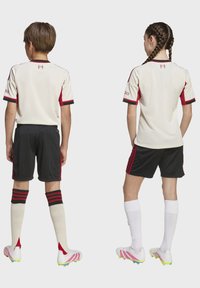 Dos niños de pie, de espaldas, vistiendo camisetas de fútbol blancas y rojas, pantalones cortos negros y zapatillas con calcetas blancas y medias negras y rojas o blancas.