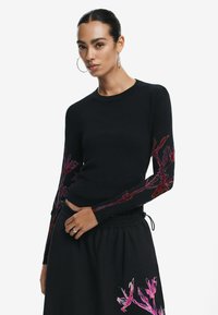 Maglione nero con maniche lunghe e ricami floreali rosa, scollo rotondo e silhouette aderente. Abbinato a una gonna nera coordinata.