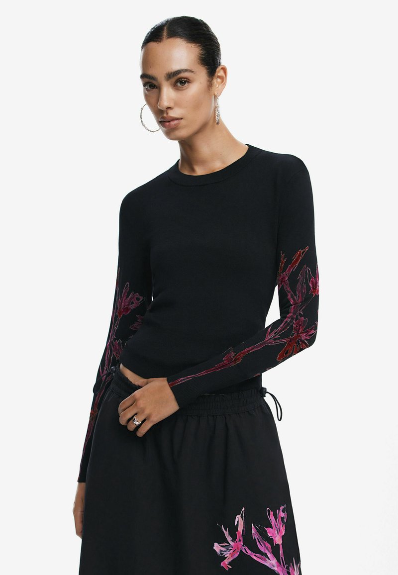Maglione nero con maniche lunghe e ricami floreali rosa, scollo rotondo e silhouette aderente. Abbinato a una gonna nera coordinata.