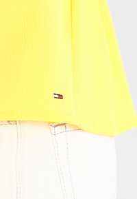 Top ligero amarillo con una superficie texturizada. Presenta un pequeño logo bordado en azul, rojo y blanco cerca del dobladillo.